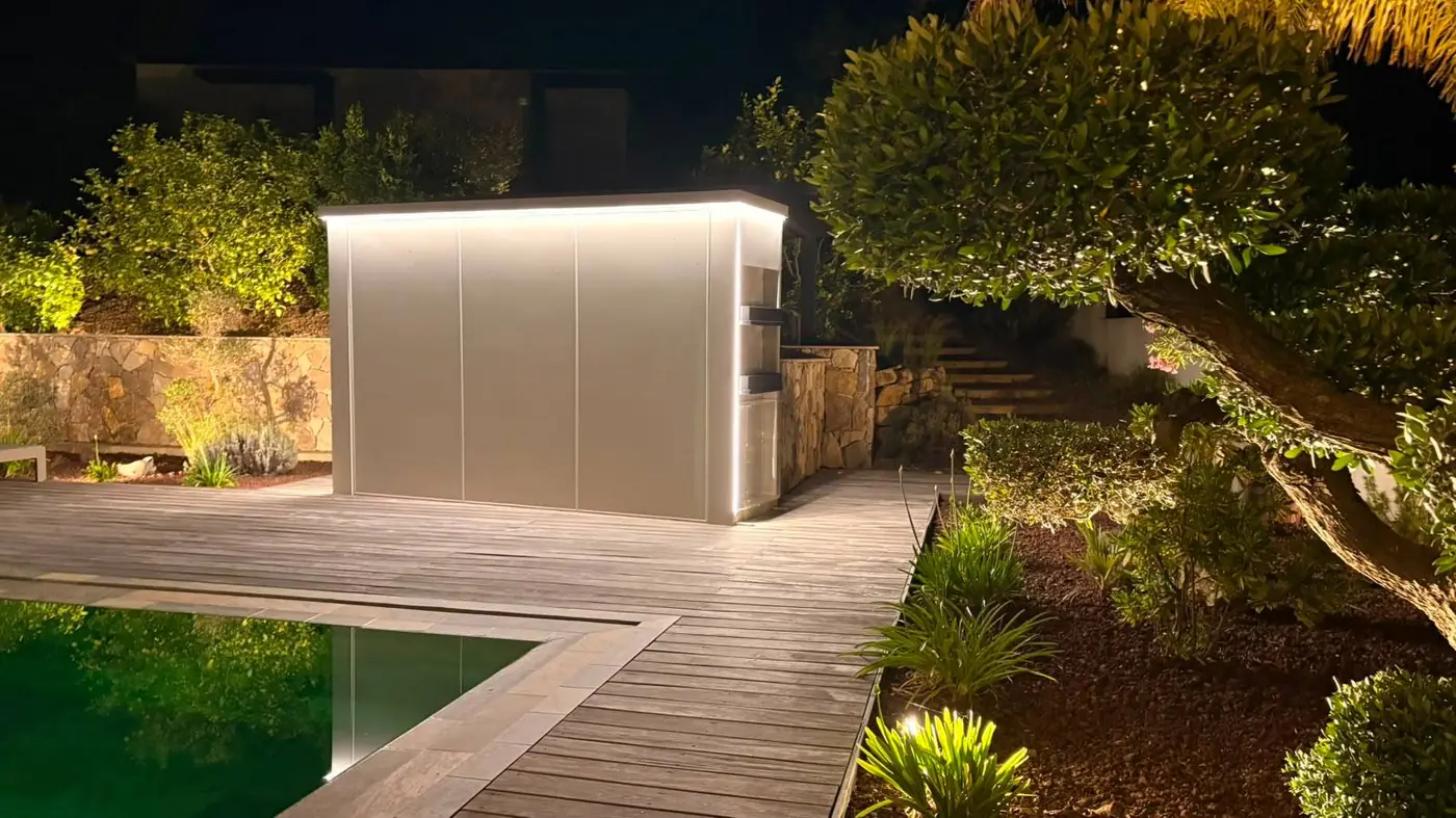 Cuisine d'extérieur Nox fermée et éclairée de nuit au bord d'une piscine sur terrasse bois