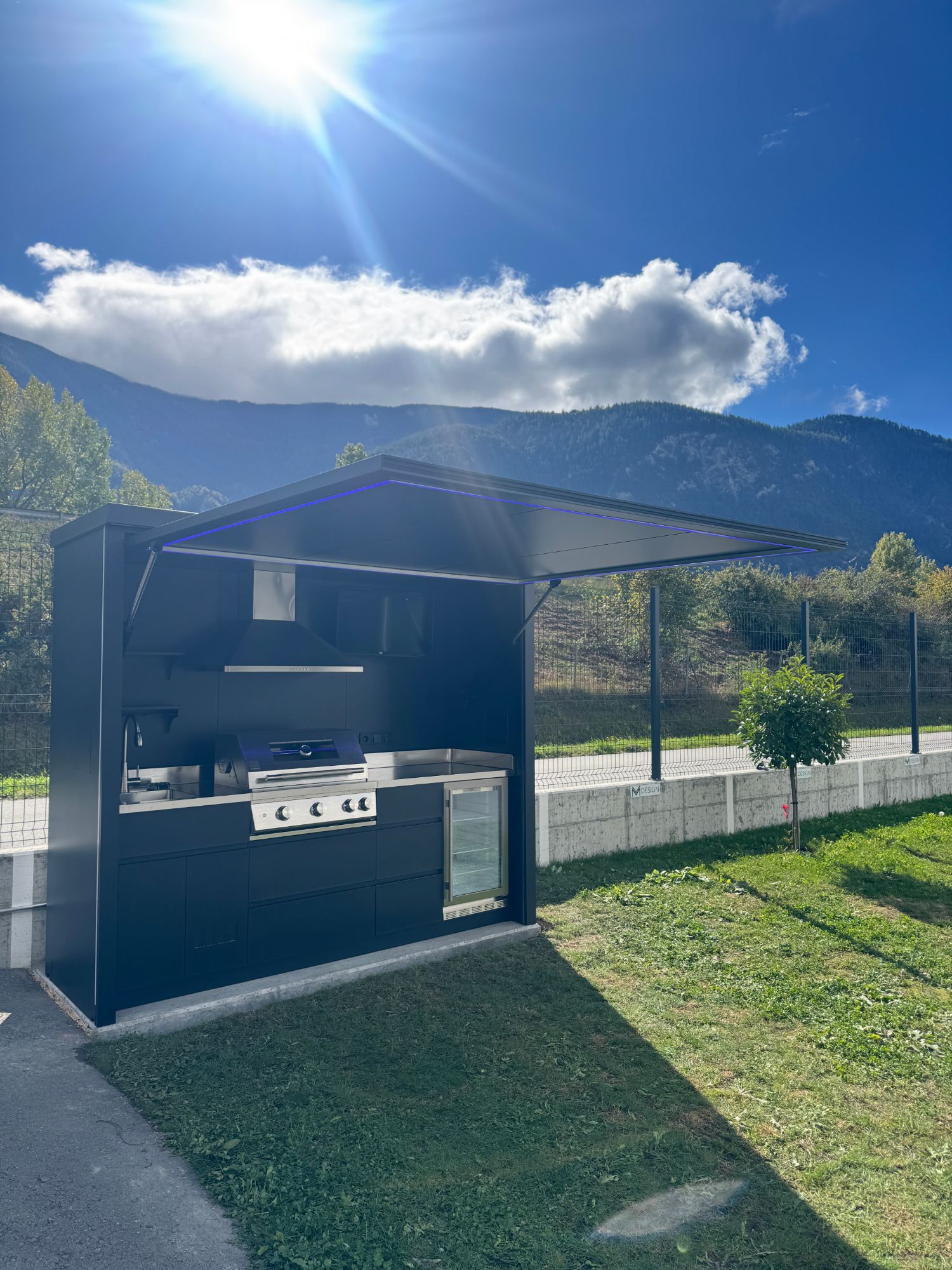 Nox Pavillon chez VM Design en Valais, vue de côté : toit ouvert avec éclairage LED bleu, évier, barbecue encastré et réfrigérateur, montagnes en arrière-plan