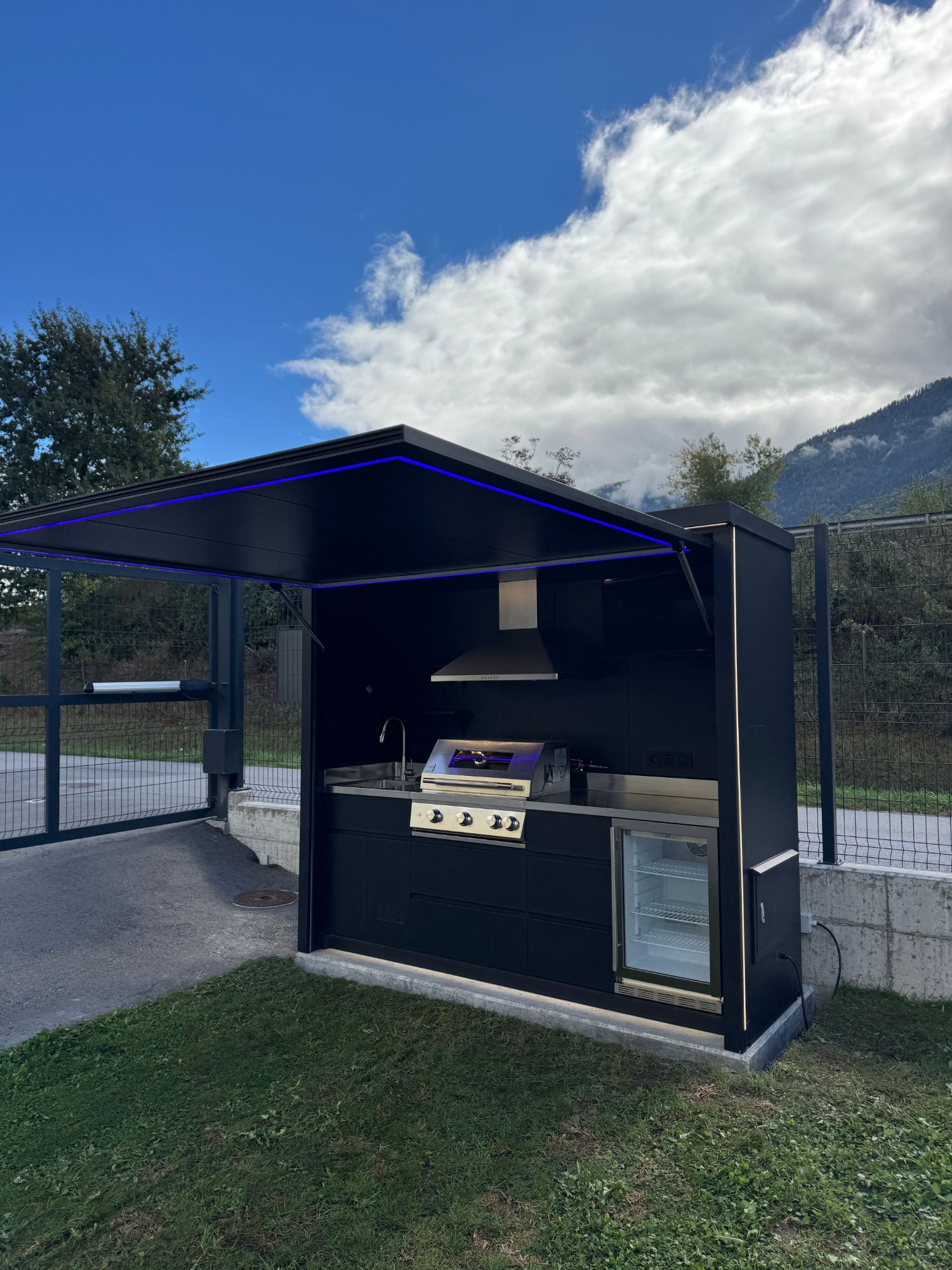 Cuisine Nox Pavillon exposée chez VM Design à Saint-Léonard, en Valais, toit relevé avec éclairage LED bleu, façade et intérieur noirs, barbecue encastré, hotte et réfrigérateur